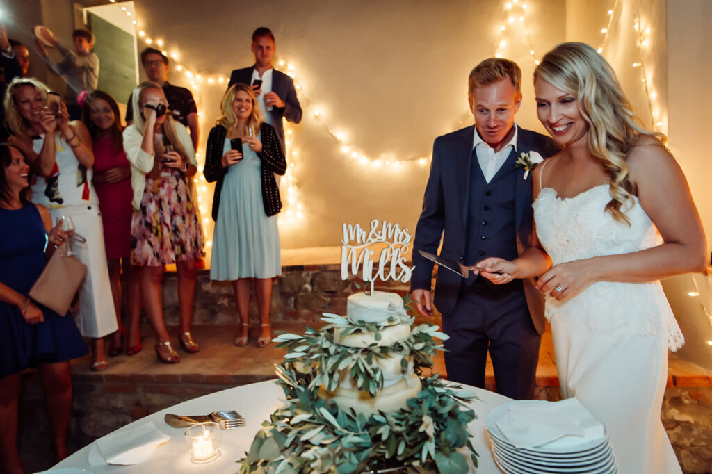 Bride and groom smiling as they start to cut the wedding cake, with guests watching and taking photos at their Tuscan wedding celebration