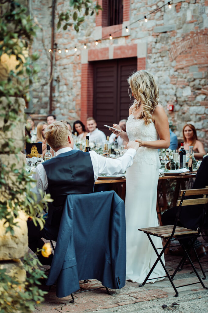 Bride giving a speech while holding hands with the groom in the castle courtyard, surrounded by guests at their Italian wedding