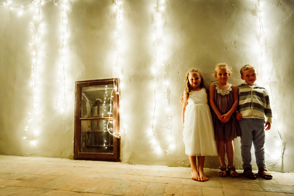 Three children standing and smiling at the camera with fairy lights behind them