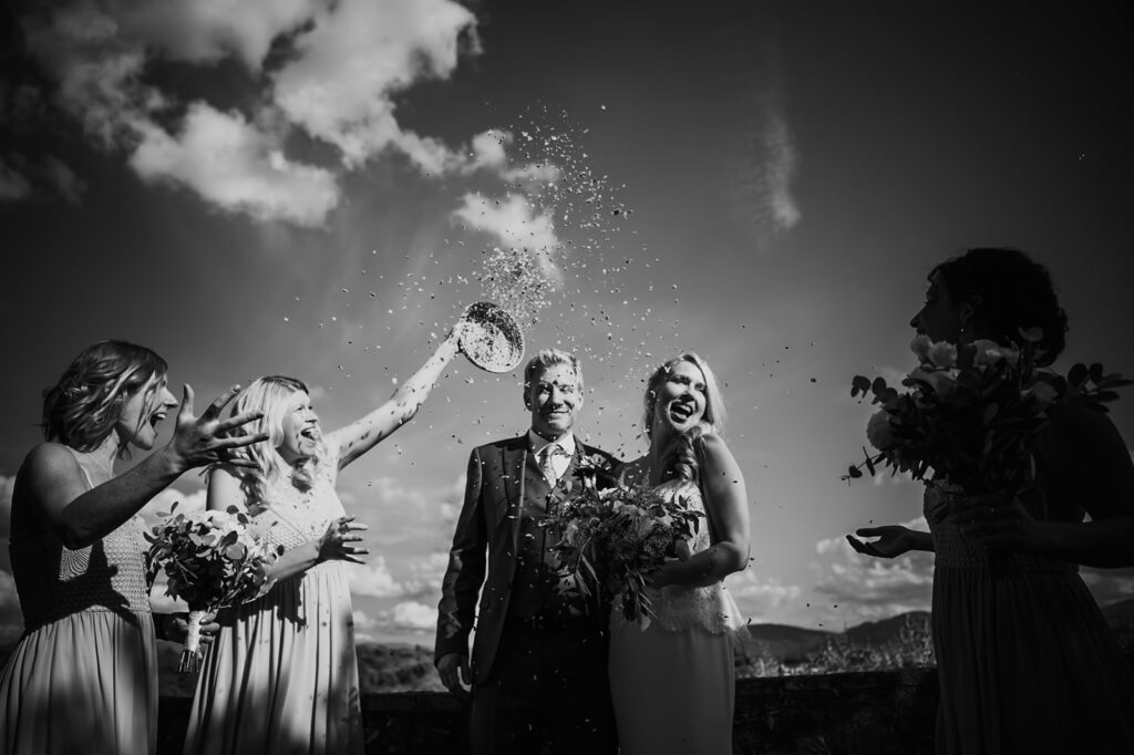 Black and white photo of newlyweds. Bridesmaids cheer and throw confetti, celebrating at Castello di Cafaggio wedding