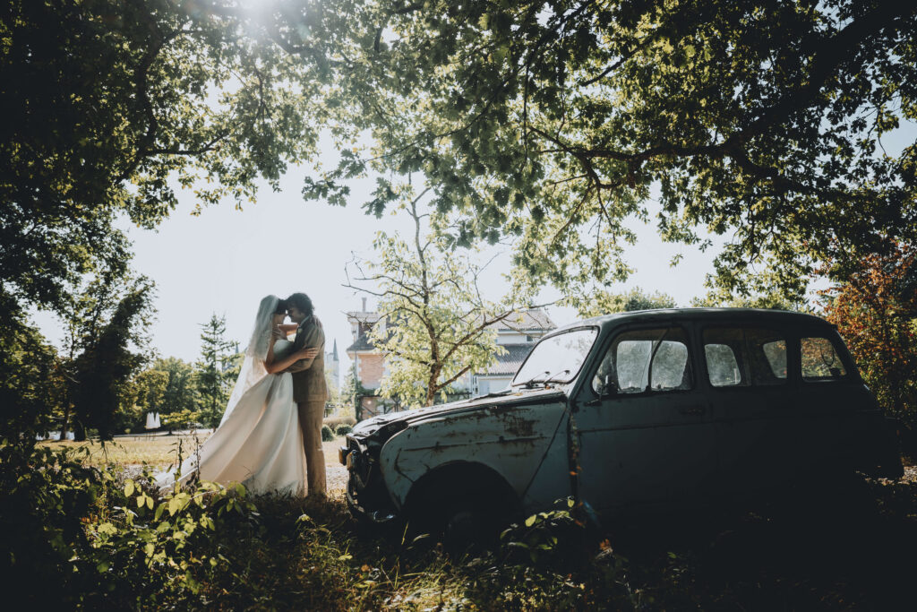 Wedding couple embracing in the shadows of surrounding trees, next to a rustic blue vintage car
