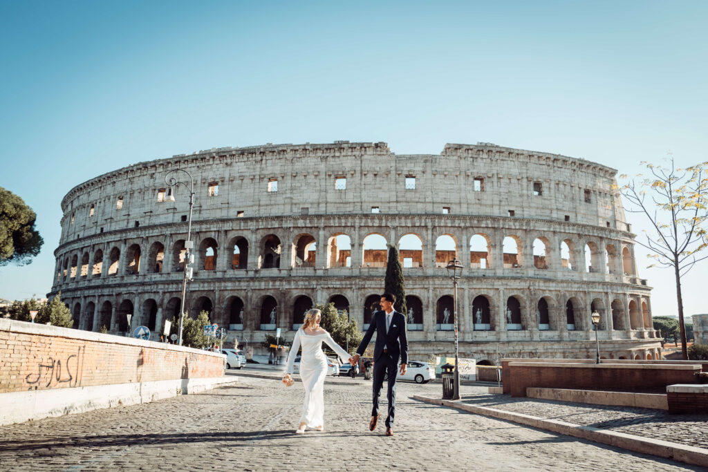 Wedding couple holding hands and walking towards the camera with the Colosseum in the background, capturing a destination wedding moment in Rome