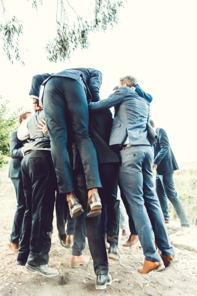 Men in suits jumping up and embracing in a joyful group moment during a wedding celebration, for 'Questions to Ask Your Destination Wedding Photographer' blog