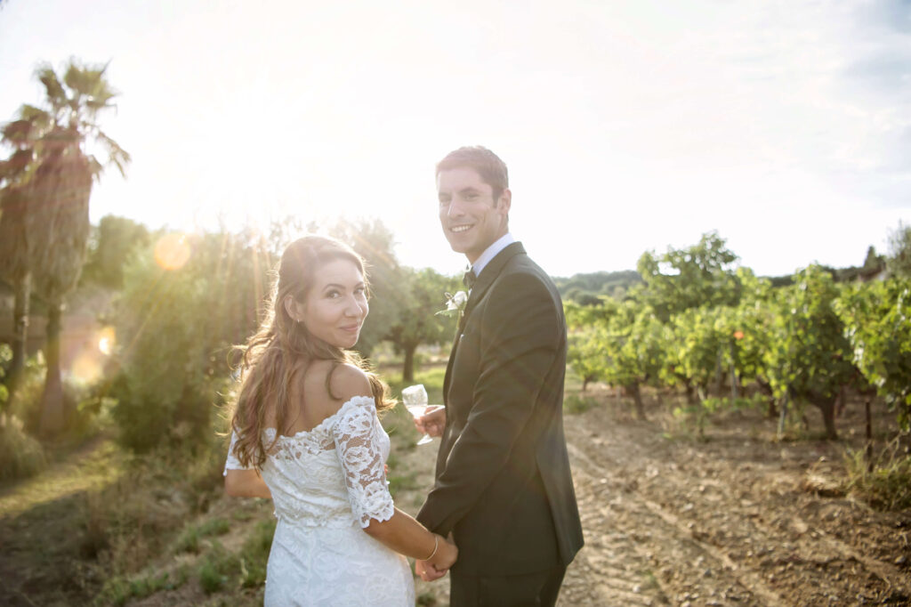 Bride and groom, holding hands, walking through a vineyard.