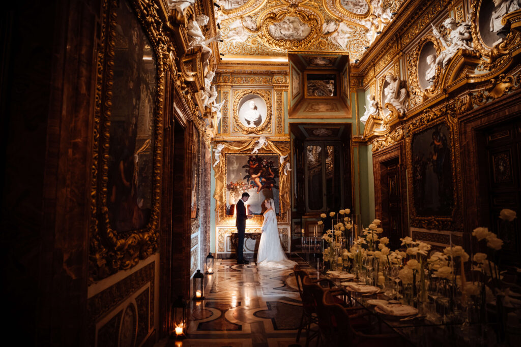 wedding couple facing each other in a grand hall with gold ceiling, ornate picture frames, and statues, moody lighting, with elegantly styled table and floral arrangements in the corner