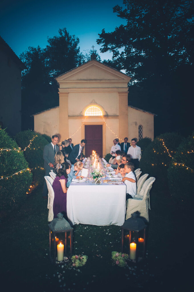 Candid shot of the wedding party getting ready for the reception meal. The sunlight is down and replaced with fairy lights and candles.