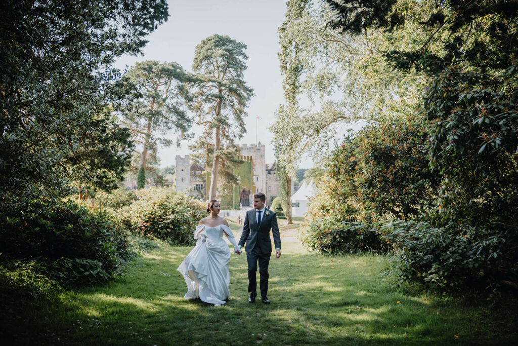 Couple holding hands walking through the Horseshoe Lawn at Hever  Castle. In the background you can see the castle through the trees.