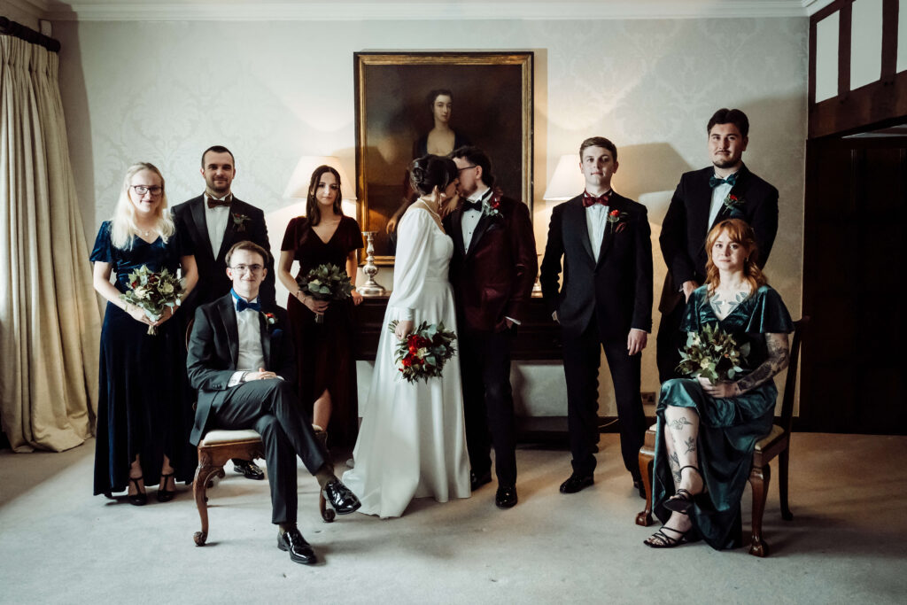 Formal shot of some wedding guests. All the guests are looking at the camera, while the couple kiss.
