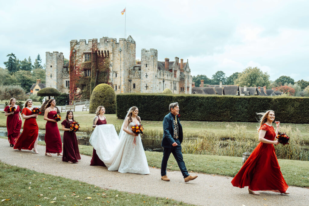 The bridal party walks together in the foreground, with the historic Hever Castle creating a timeless backdrop behind them