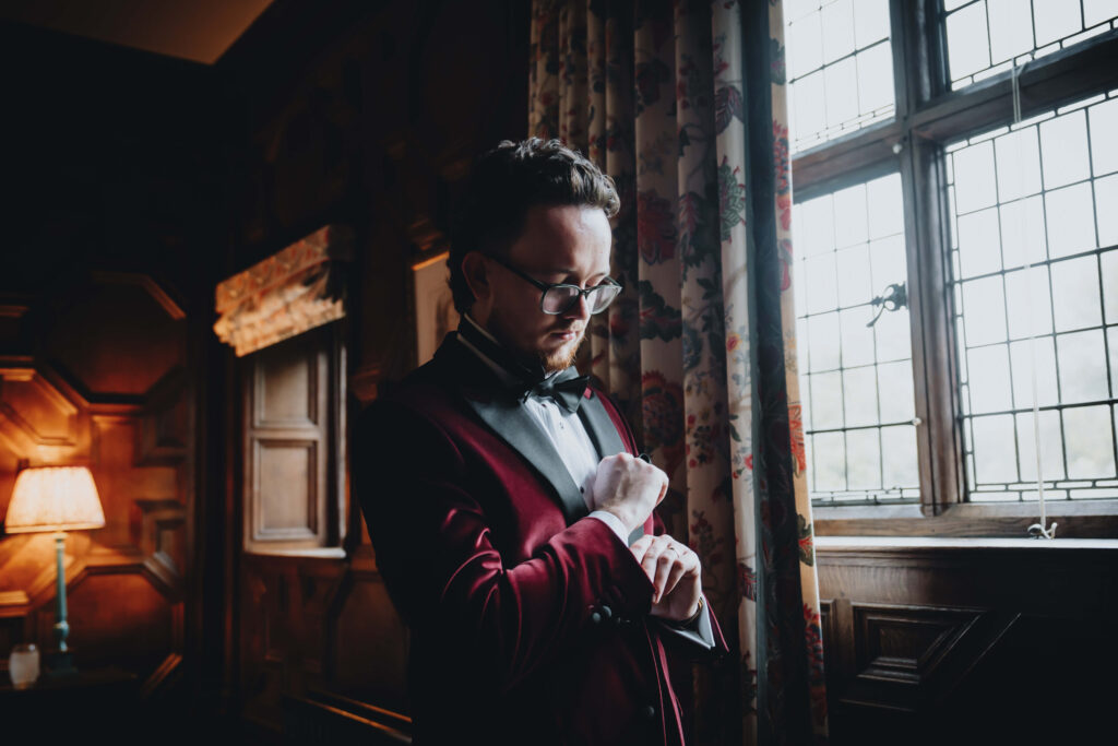 Groom standing next to a window, adjusting his cuffs