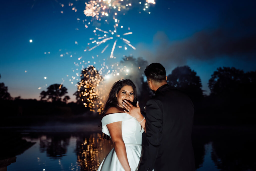 The background shows a late evening with fireworks. In the fore grand a wedding couple hold hands. The groom watches the fireworks while the bride looks back at the camera.