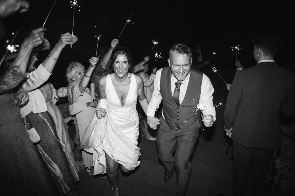 Black and white photo of couple running through wedding guests, who are holding sparklers.