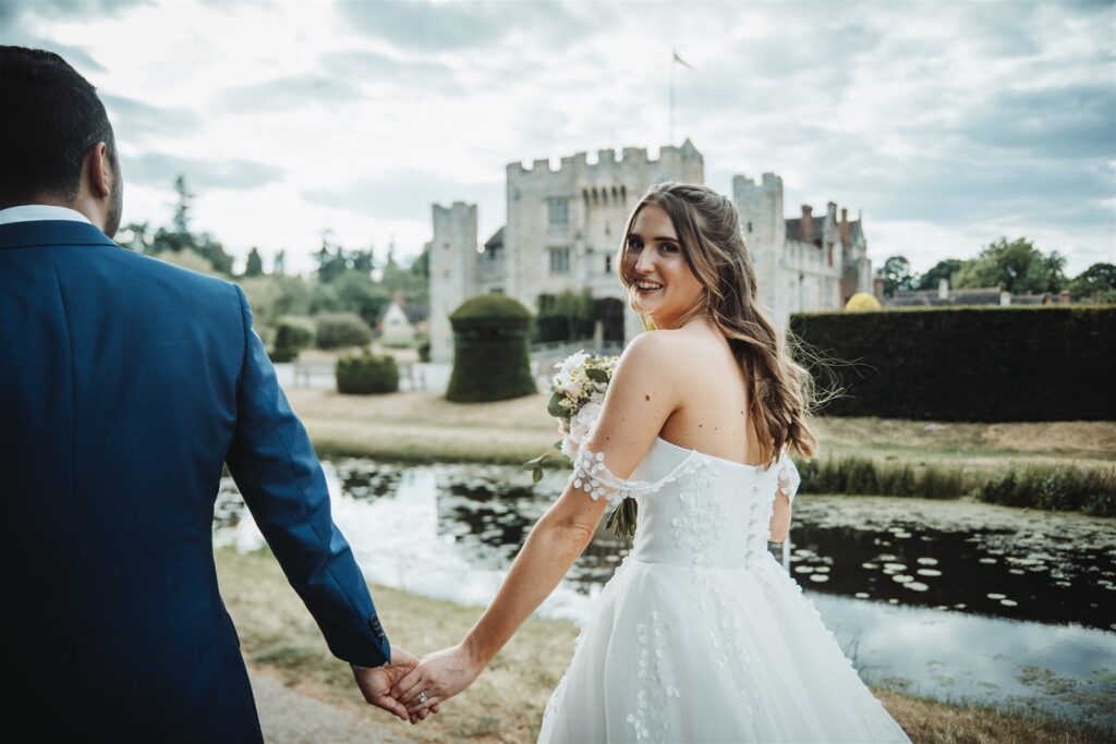 Bride and groom holding hands with the bride looking over her shoulder and smiling, with Hever Castle in the background, captured by a Hever Castle wedding photographer