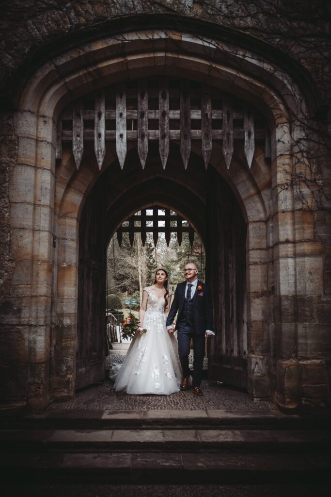 Bride and groom walking hand in hand through an old castle gate with a raised portcullis