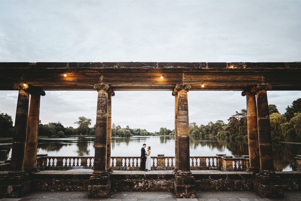 Wide shot of a couple standing between four pillars by the lake at Hever Castle
