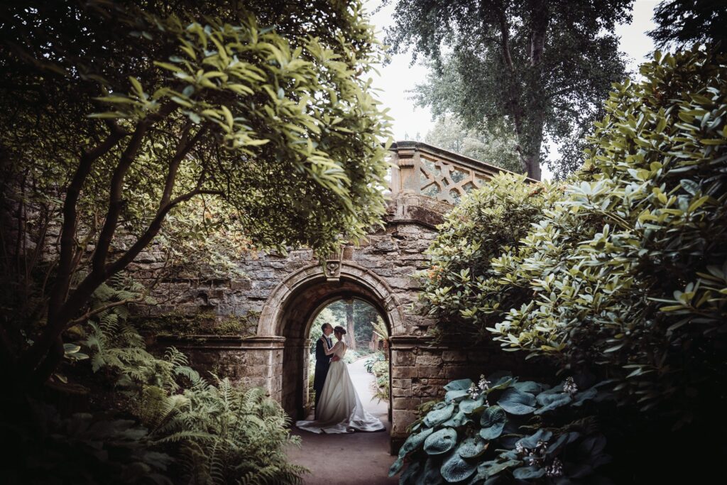 Bride and groom sharing an intimate moment beneath a historic stone archway surrounded by lush greenery