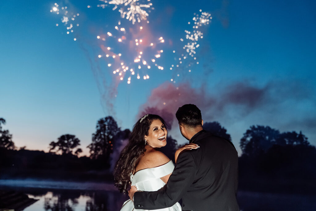 Wide shot of a couple from behind watching fireworks at Hever Castle, the bride looking back and smiling