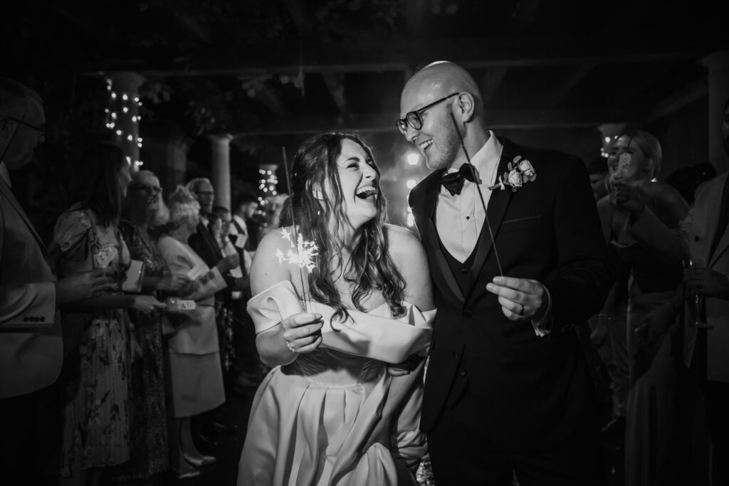 Black and white candid shot of a couple laughing and holding sparklers, surrounded by their wedding party at Hever Castle