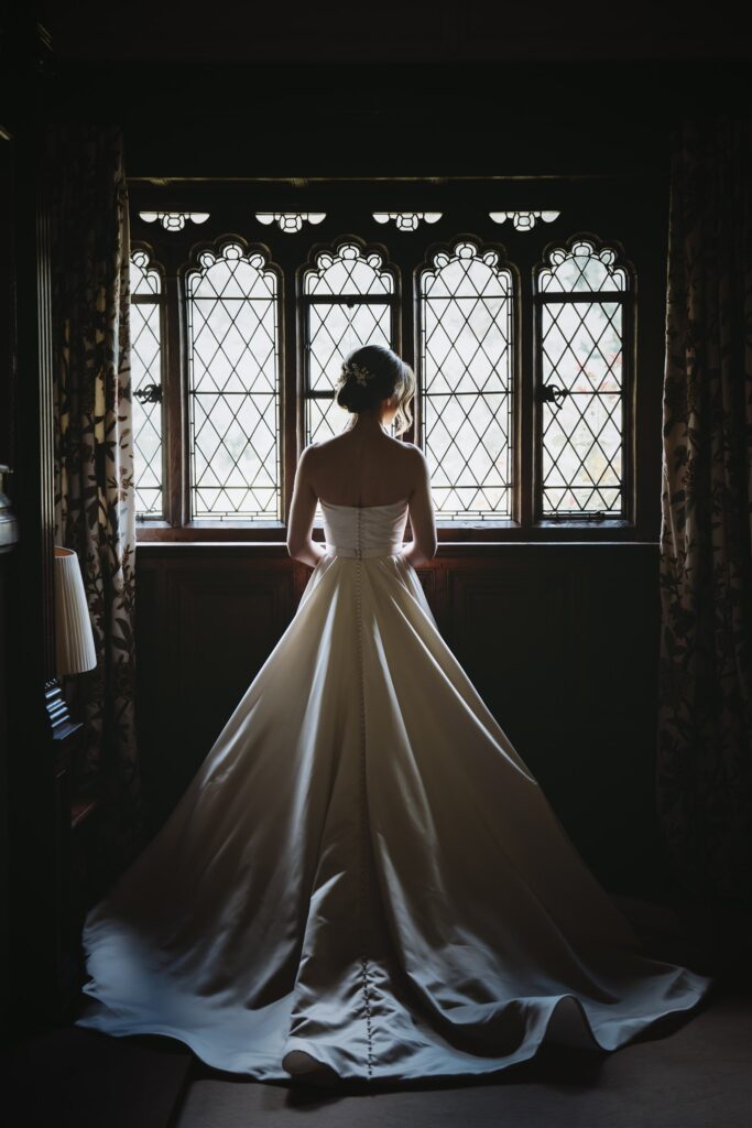 Bride standing inside Hever Castle looking out a window, showing the back of her wedding dress, in dark lighting. Photographed by a Hever Castle wedding photographer.