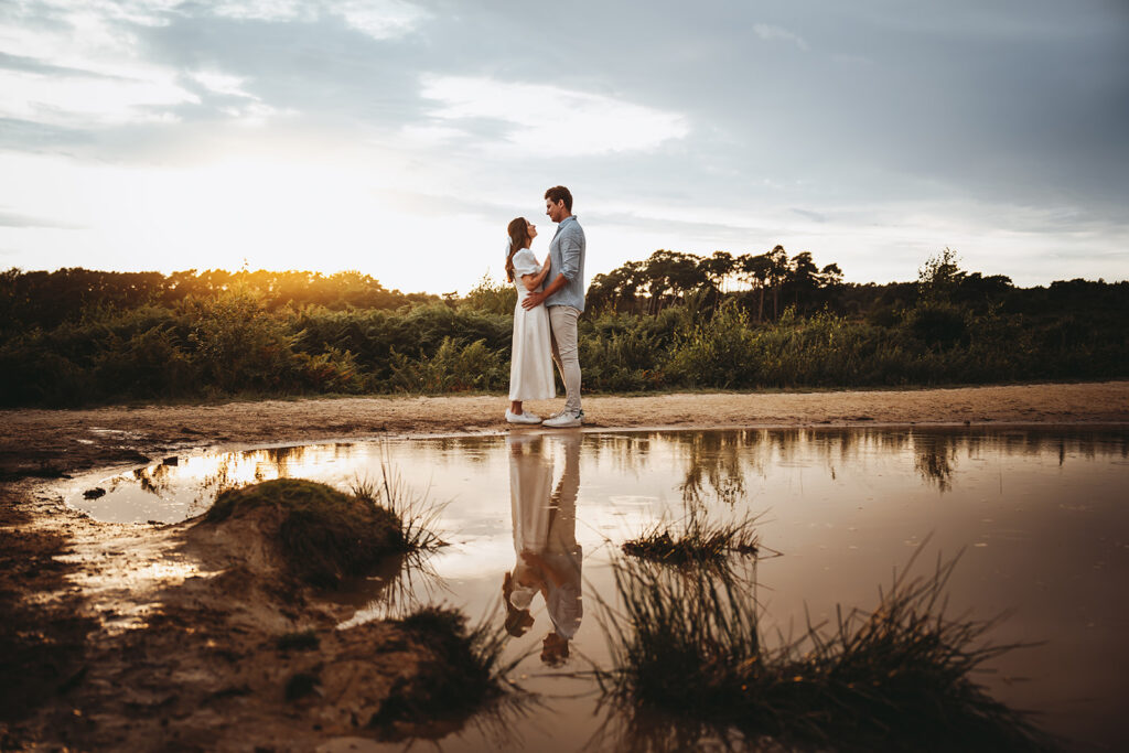 Couple embracing and looking at each other next to a lake with a small sunset behind them. [ A Wedding Timeline Guide ]
