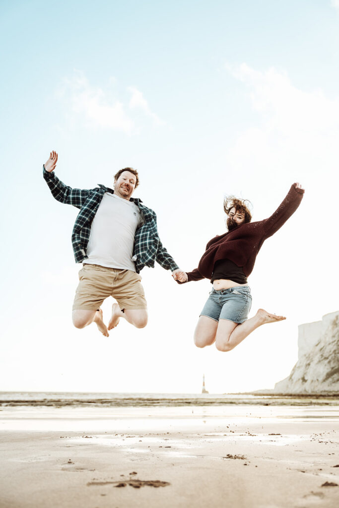 Couple holding hands and jumping in sync on a beach near cliffs, with a lighthouse in the distance. [Teri V Photography Wedding Timeline Guide]